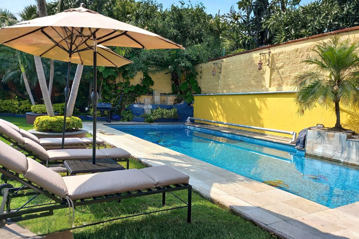 Área exterior en Las Fuentes 34-A con alberca, camastros sobre césped y muro amarillo decorativo.