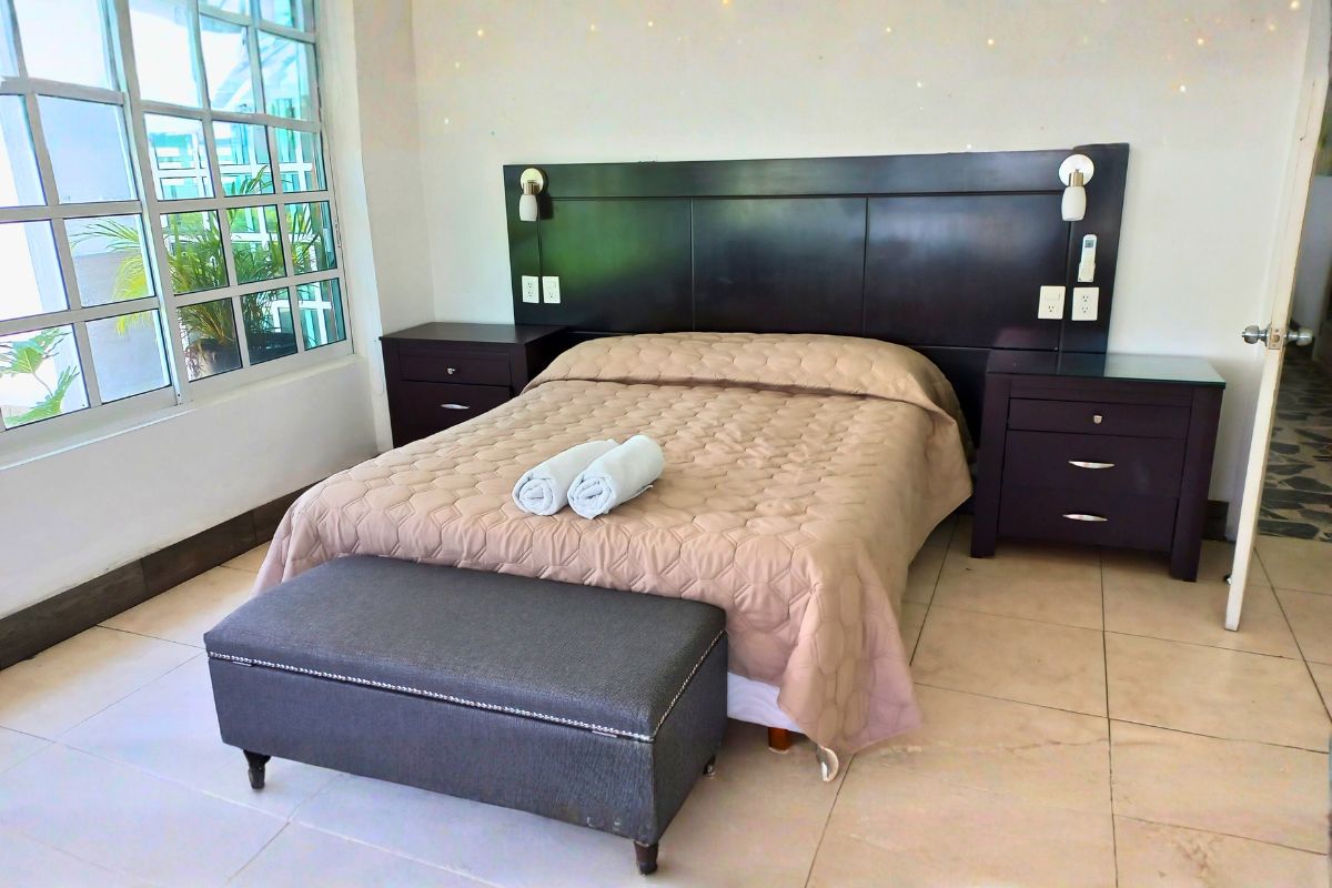Recámara principal en Pedregal 20 con cama queen, cabecera de madera, bancas y luz natural.