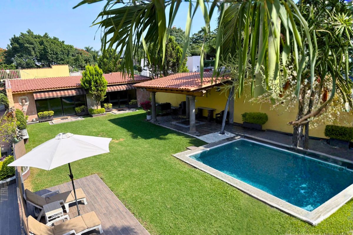 Área exterior en Villa Las Fuentes con alberca, terraza techada, camastros y vegetación tropical.